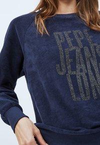Femme portant un sweat-shirt bleu marine avec le texte scintillant "PEPE JEANS LONDON", ajustant l'ourlet d'une main.