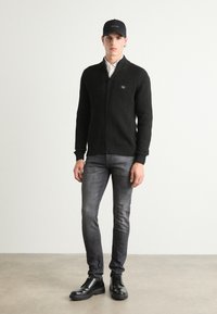 Maglione nero a coste con zip e colletto alto, jeans grigi slim-fit e stivali neri con lacci, accompagnati da un berretto nero con logo bianco.