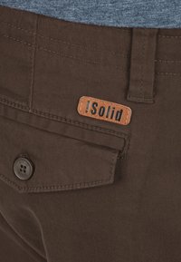 Un pantalon en tissu brun présente une surface texturée avec une poche arrière, un bouton noir et une étiquette en cuir beige portant la mention "!Solid."