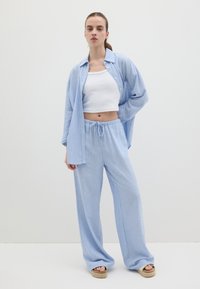 Camicia oversize a strisce azzurre con maniche lunghe, top corto bianco e pantaloni azzurri con cordoncino abbinati. Sandali con tacco completano il look.
