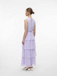 Sieviete ar sasietu matu kušķi, kas valkā piedurkņu neesošu lavandas krāsas daudzslāņu maxi kleitu un baltus blokpapēžu apavus, stāv sāniski pret balto fonu.