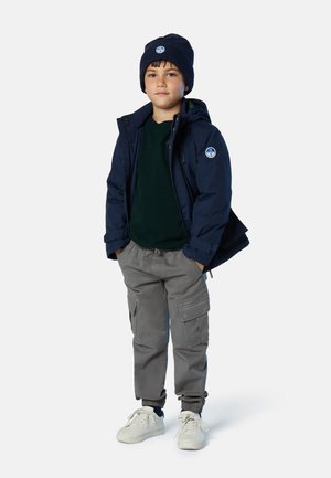 Chaqueta acolchada azul marino con parche de logo, suéter verde, pantalones cargo grises y zapatillas blancas. El gorro combina con el color de la chaqueta.