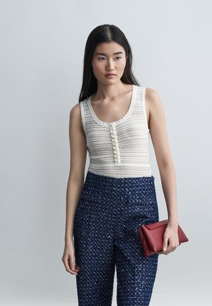 Jeune femme aux longs cheveux noirs portant un haut blanc sans manches, un pantalon bleu marine texturé, et tenant une pochette rouge.