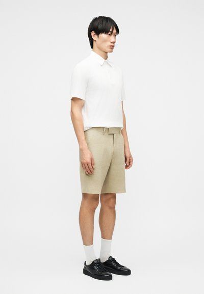 Νεαρός άνδρας που στέκεται φορώντας λευκό πόλο με κοντά μανίκια, μπεζ σορτς με ζώνη, λευκές κάλτσες και μαύρα αθλητικά παπούτσια σε λευκό φόντο.