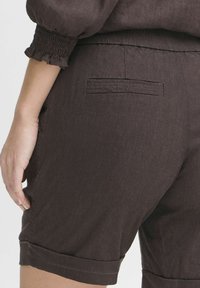 Brown linen shorts with rolled cuffs, featuring a back pocket and a textured fabric. The waistband is elasticated for comfort.