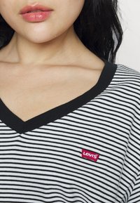 Černobílý pruhovaný tričko s v-výstřihem a červenou štítkou Levi's na levé straně. Tkanina vypadá hladce a je lehká.