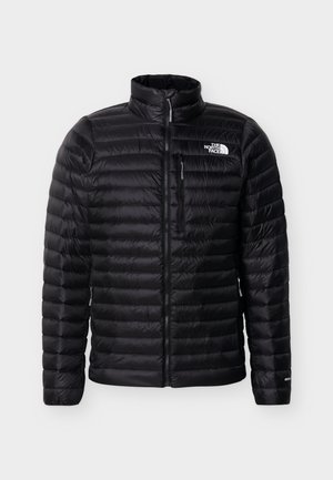Musta tikattu North Face -takki, jossa on koko pituinen vetoketju edessä, rintataskun vetoketju ja logo vasemmassa ylärinnassa, valkoista taustaa vasten.
