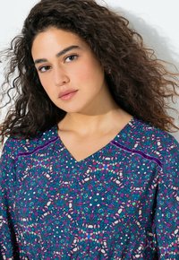Junge Frau mit lockigem braunem Haar, die ein farbenfrohes, gemustertes Oberteil mit lila, blauen und rosa abstrakten Formen vor einem weißen Hintergrund trägt.
