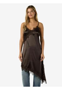 Sieviete ar gariem blondiem matiem, valkājot tumšu sat�īna naktskrekli ar mežģīņu apdari pār gaiši zilām džinsu biksēm, stāvot pret vienkāršu baltu fonu.