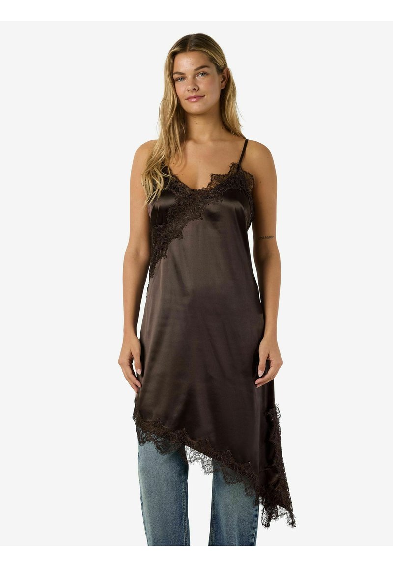 Sieviete ar gariem blondiem matiem, valkājot tumšu satīna naktskrekli ar mežģīņu apdari pār gaiši zilām džinsu biksēm, stāvot pret vienkāršu baltu fonu.