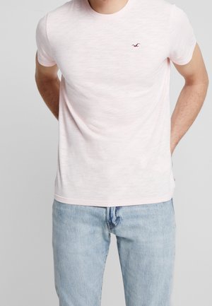 Jasnoróżowa koszulka z krótkim rękawem, uszyta z miękkiego, lekkiego materiału, z małym ciemnym logo na klatce piersiowej i luźnym krojem.