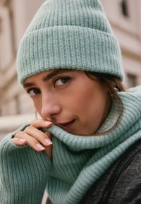 Light green, ribbed knit beanie and matching scarf, showcasing soft texture. Close-up view reveals delicate fingers holding the scarf.