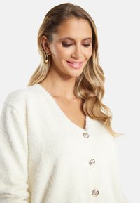 Bílý, nadýchaný cardigan s hlubokým výstřihem ve tvaru V a čtyřmi lesklými knoflíky na zapínání, se soft texturou a bez viditelných vzorů nebo akcentů.