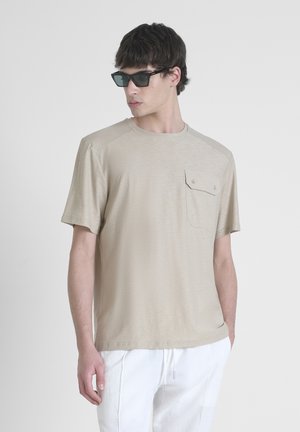 Camisa de manga corta beige con un acabado texturizado, que presenta un bolsillo en el pecho con detalles de botones. Combinada con pantalones de lino blancos y gafas de sol.