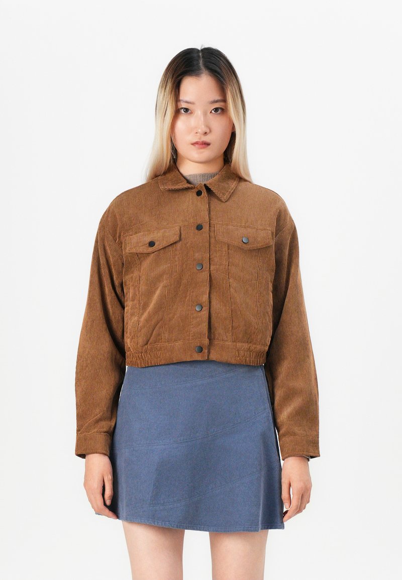 Jeune femme avec des cheveux raides ombrés, portant une veste en velours côtelé marron courte et une mini-jupe bleue, se tenant contre un fond blanc uni.