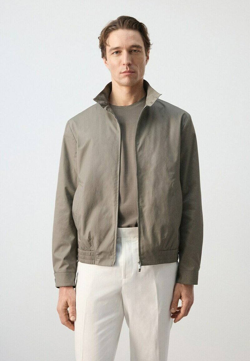 Moški, oblečen v svetlo rjavo jopo z zadrgo, taupe majico in bele hlače, stoji pred enobarvnim svetlo sivim ozadjem.