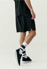 Fekete sportnadrág oldalsó hasítékokkal, fekete cipővel és fekete csíkkal díszített fehér zoknival párosítva. Simó, könnyű anyag.