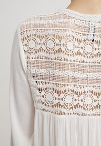 Vit blus med en texturerad, spetsbakpanel med cirkulära mönster. Tyget är mjukt och har veck nedanför oket.