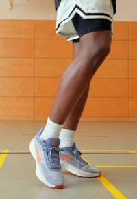 Lysegrå sportssko med orange detaljer, mesh-overdel og gummisål. Båret med hvide ankelstrømper og sorte sportsshorts på et fitnessgulv.