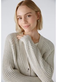 Hellgrauer Strickcardigan mit geripptem Muster, offener Vorderseite und einem einzelnen Knopfverschluss. Verfügt über lange Ärmel und eine lockere Passform.