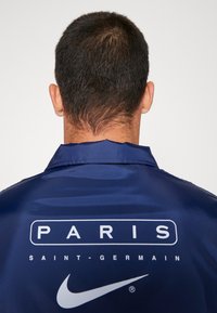 Navy satin jacket featuring a white printed logo reading "PARIS SAINT-GERMAIN" and Nike's swoosh on the back. Smooth texture and collar design.