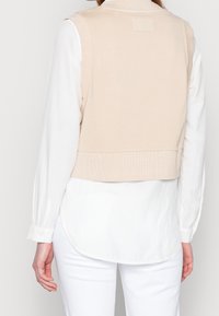 Chaleco de punto beige recortado con textura acanalada, cuello alto y parche rectangular en la parte trasera, usado sobre una camisa blanca de manga larga.