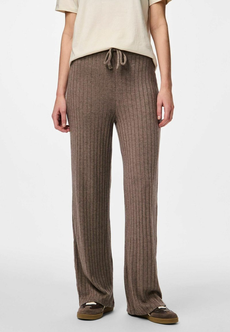 Personne porte un pantalon en maille marron ample et côtelé avec une taille à cordon et des chaussures marron foncé à enfiler, assorti à un haut beige à manches courtes.