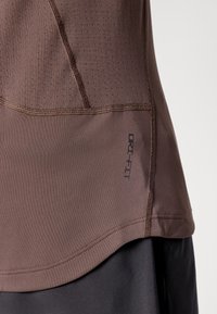 Nærbilde av en brun treningstrøye med "DRI-FIT"-tekst på sidesømmen, over svarte bukser, som viser detaljert søm og tekstur i stoffet.