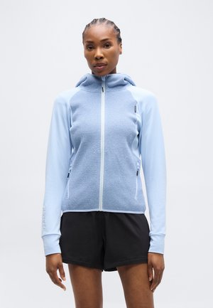 Fonott hajú nő világoskék cipzáras kapucnis felsőben, melynek elöl texturált paneljei vannak, fekete sportnadrágban, egy egyszínű háttér előtt állva.