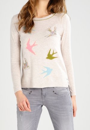 Kvinde iført en lys beige langærmet sweater med flerfarvede fuglemønstre og falmede grå jeans, stående mod en ensfarvet baggrund.