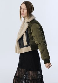 Frau in olivgrüner Bomberjacke mit beigem Lammfellbesatz über einem transparenten schwarzen Spitzenrock, die seitlich vor einem einfarbigen hellen Hintergrund posiert.