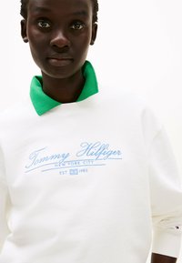 Witte sweatshirt met een groene kraag. Bevat blauwe geborduurde tekst "Tommy Hilfiger New York City EST 1985." Zachte textuur, casual ontwerp.
