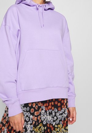 Personne portant un sweat à capuche violet clair avec une poche avant et une jupe noire à motifs de petites fleurs multicolores.