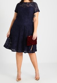 Navy lace A-line dress with short sleeves, floral pattern, and a flared skirt. Paired with a burgundy velvet clutch and nude heels.