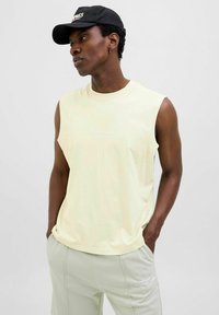 T-shirt sans manches en coton jaune clair avec encolure ronde et logo "Originals Studio" sur la poitrine, associé à un pantalon gris clair.