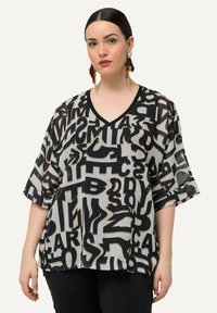 Sort-hvid mønstret transparent bluse med V-udskæring og løse ærmer, dekoreret med store abstrakte bogstaver, der dækker stoffet.