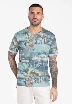 Homme aux cheveux courts et tatouages portant un t-shirt graphique bleu représentant des voitures anciennes, des palmiers, des planches de surf et des surfeurs dans un paysage urbain côtier.