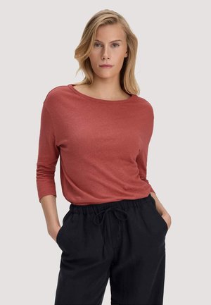 Blonde vrouw met een roestkleurig shirt met lange mouwen en zwarte broek met trekkoord, staand met handen in de zakken tegen een lichte achtergrond.
