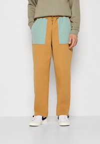 Braune Sweatpants mit hellblauen strukturierten Taschen, elastischem Bund mit Kordelzug, kombiniert mit weißen Sneakers mit schwarzen Akzenten.