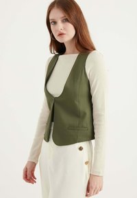 Olivgrön väst med V-hals, utan knappar, slim fit och två framfickor, bärs över en krämfärgad långärmad topp och vita byxor.