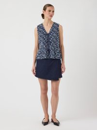 Blouse sans manches bleu marine avec un motif floral, ourlet peplum et détail de boutons, associée à une jupe portefeuille en denim foncé et des ballerines noires pointues.
