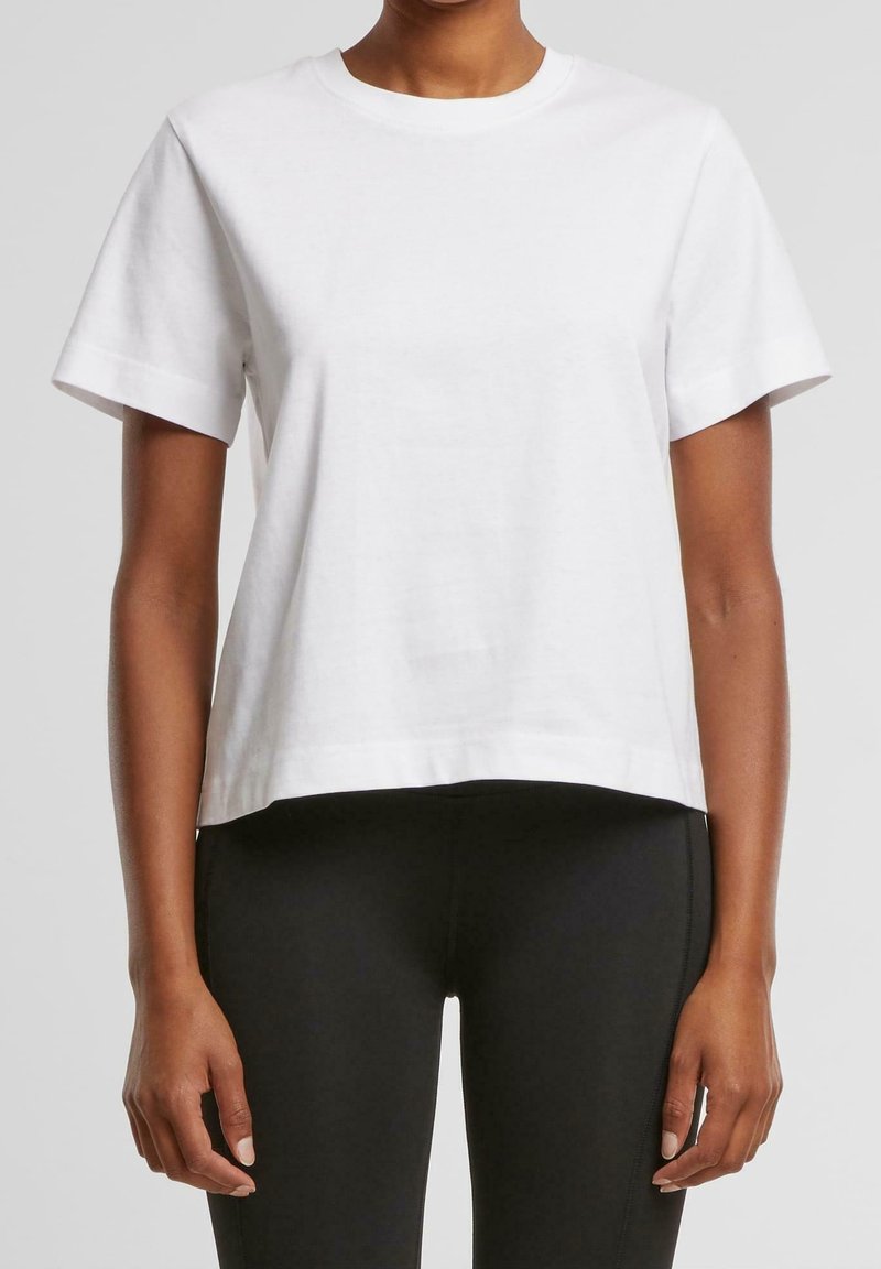 Personne portant un t-shirt blanc uni à manches courtes légèrement rentré dans un pantalon noir ajusté, visible du cou jusqu'aux hanches sur un fond neutre.