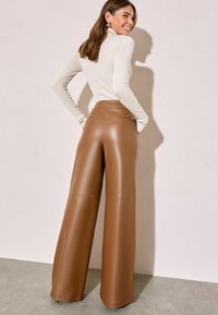 Pruuni kunstnahast laiad püksid kõrge v�öökohaga, siledat tekstuuri ja peene õmbluse detailidega. Mudel kannab heledat istuvat topi.
