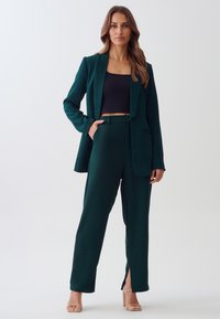 Tmavě zelený oblek se padnoucím sakem, odpovídajícími kalhotami s předními rozparky a černým crop topem. Hladká textilie, minimalistický design.