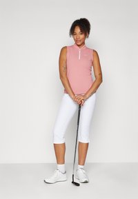 Top sin mangas de color rosa claro con un cuello de media cremallera, combinado con pantalones capri blancos. El modelo sostiene un palo de golf y lleva zapatos blancos.
