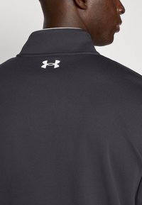 Σκούρο γκρι πόλο πουκάμισο με υφασμάτινη υφή, με λευκό λογότυπο Under Armour στο πίσω μέρος του  γιακά. Απλός σχεδιασμός με όρθιο γιακά.