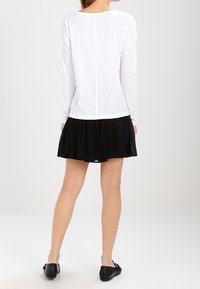 Kvinna modell sedd bakifrån, iförd en vit långärmad topp och en svart utsvängd kjol, stående mot en ljus bakgrund.