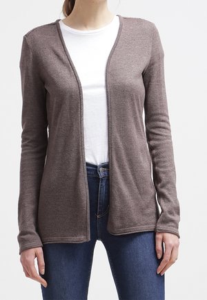 Personne portant un cardigan marron ouvert devant, sur une chemise blanche et un jean bleu, debout devant un fond clair uni.