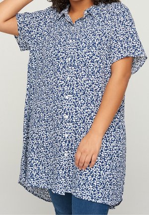Kobieta nosząca luźną, krótką tunikę z guzikami w niebiesko-biały kwiatowy wzór na niebieskich dżinsach.