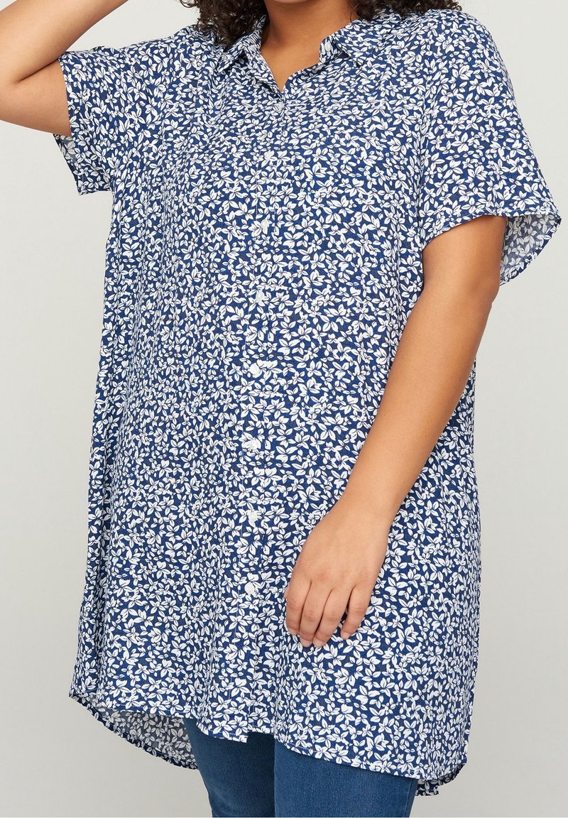 Femme portant une tunique ample à manches courtes boutonnée avec un motif floral bleu et blanc sur un jean bleu.